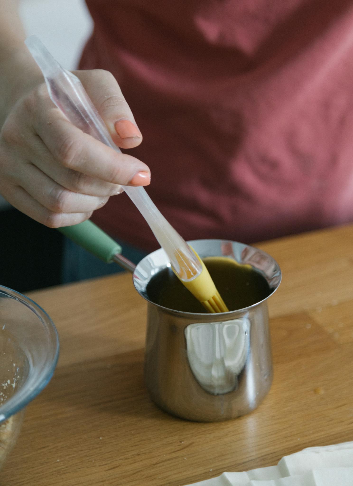 Close-up of a hand using a brush to apply oil in a stainless steel container indoors.
